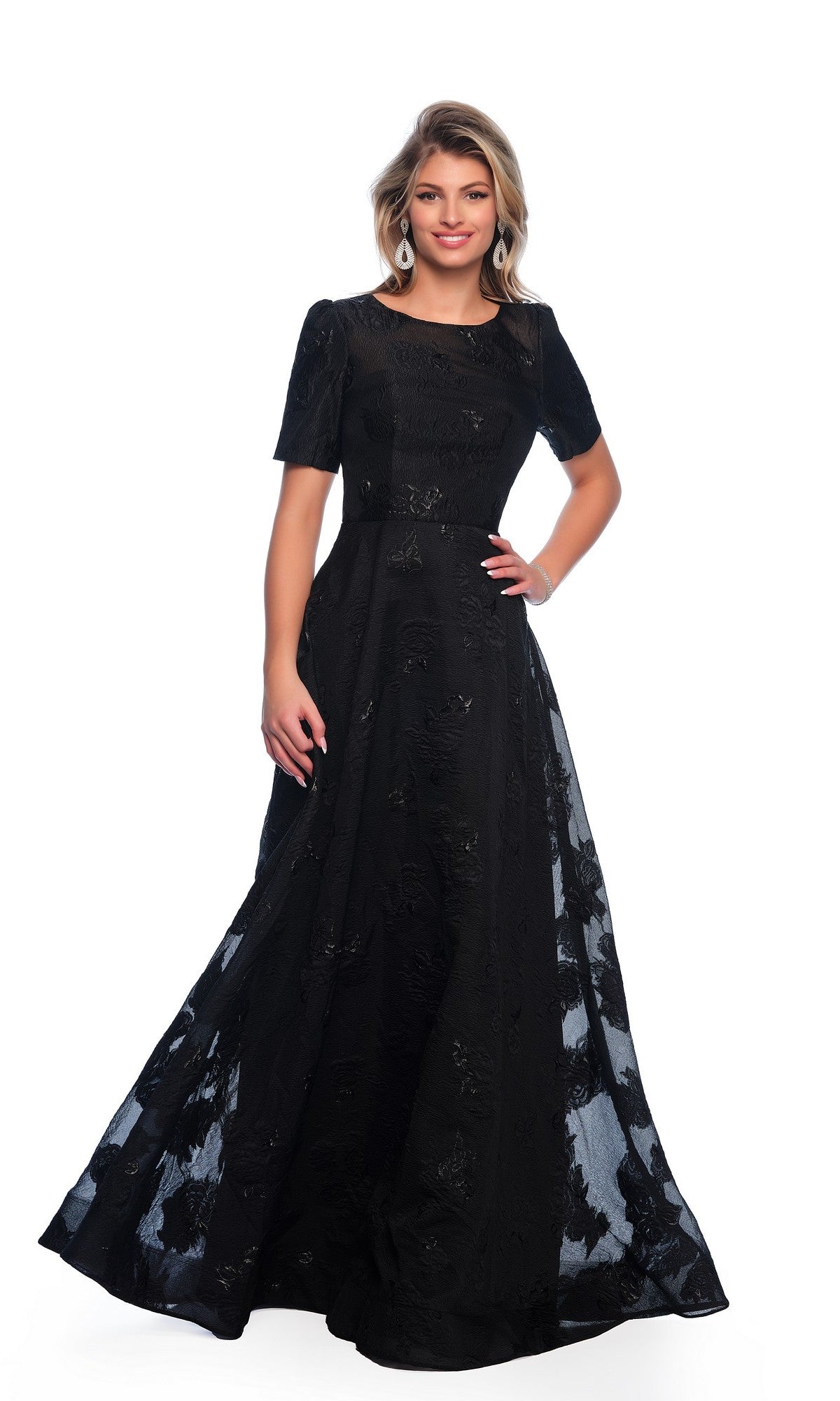 A woman stands against a plain white background wearing the Modest Black Prom Dress: Dave and Johnny 11815, a floor-length gown with short sleeves and a subtle floral pattern.